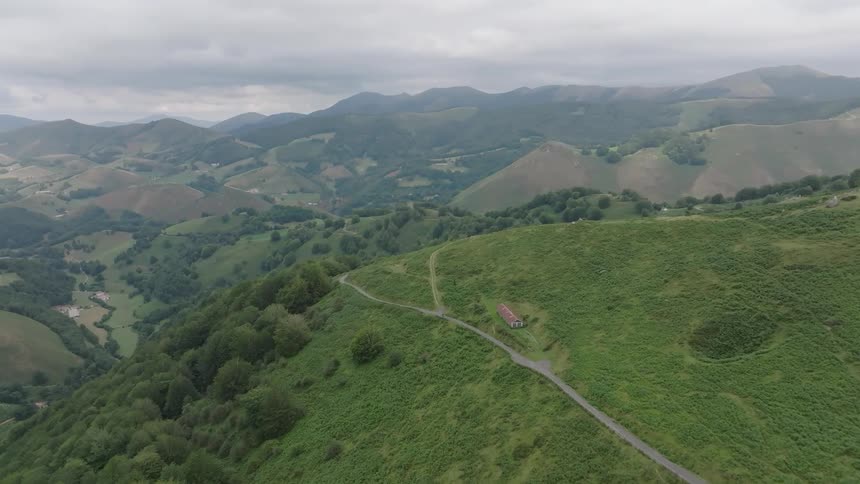 A breathtaking aerial shot slowly reveals a lonely road winding its way through endless lush green hills and valleys on a cloudy day
