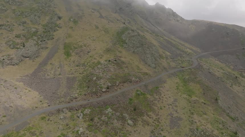 An epic aerial view follows a lone cyclist riding along a winding dirt road through a vast and foggy mountain range