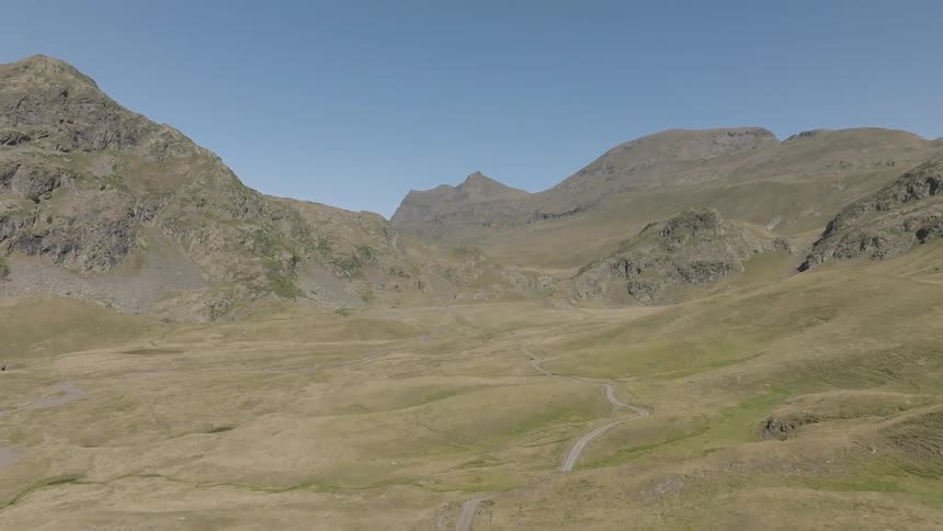 An aerial view slowly advances over a vast and rugged mountain valley, revealing a small tarn and a winding dirt road on a clear summer day