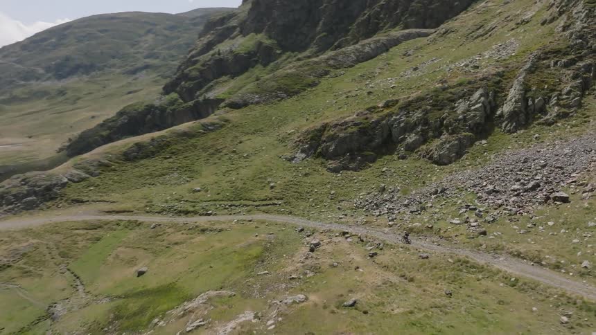 An aerial shot follows a lone mountain biker riding on a gravel path through a vast and beautiful green valley