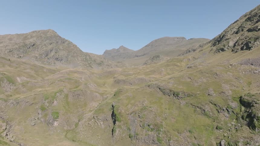 A stunning aerial view flies through a vast, rugged mountain valley with a meandering stream on a clear summer day