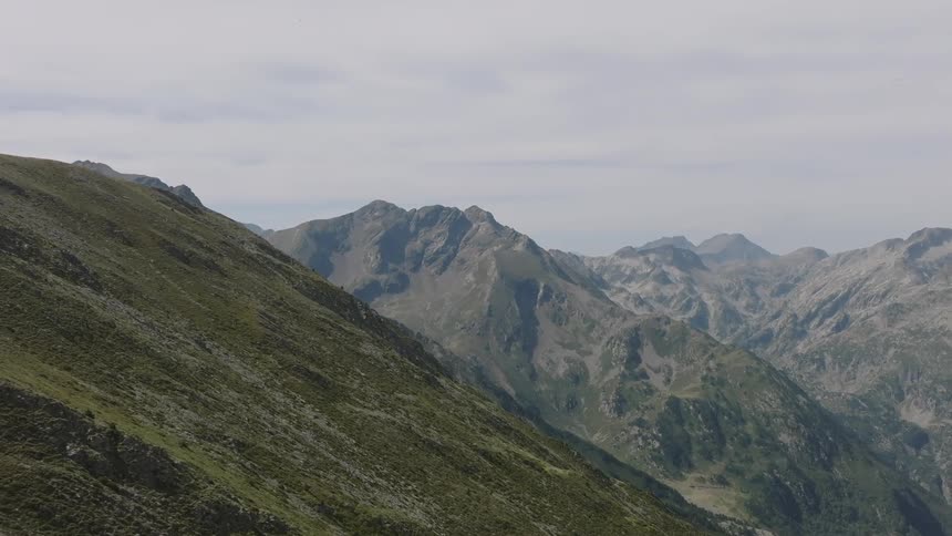 A sweeping aerial view pulls back over a steep, green mountain slope to reveal a vast, rugged mountain range