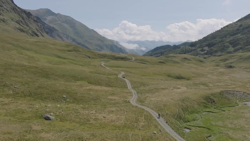An epic aerial shot follows a lone mountain biker riding along a winding dirt road through a vast, green alpine valley