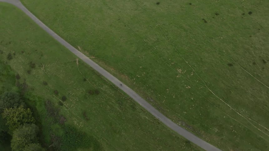 An aerial top-down view shows a lone cyclist riding along a narrow country road through a vast green field