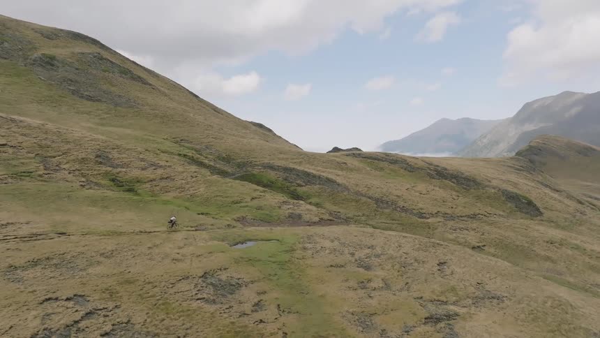 An aerial shot follows a lone mountain biker traversing a scenic trail across a vast, grassy mountainside
