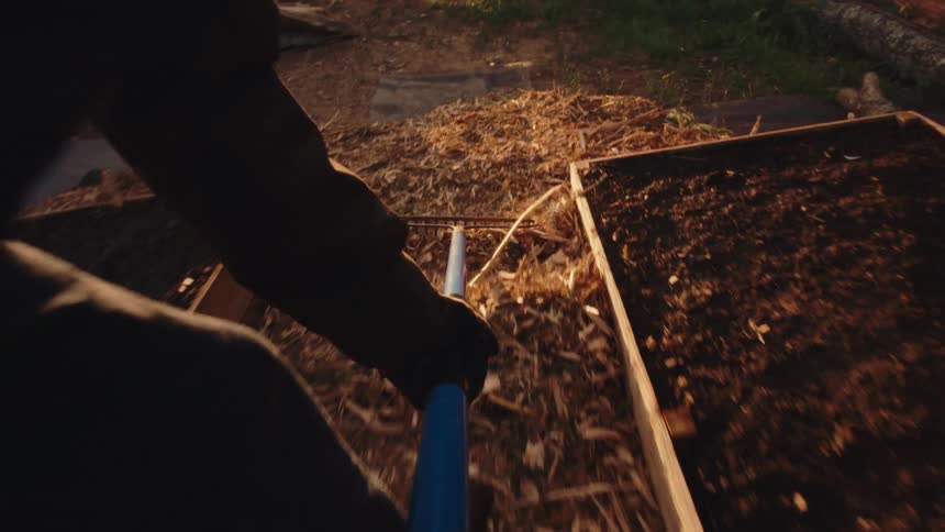A stylized, dizzying first-person view of a person raking mulch around garden beds at golden hour