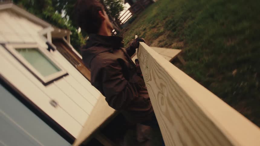 A young man carries a long wooden plank toward a small house, working on a building project in this slow-motion, stylized vertical shot