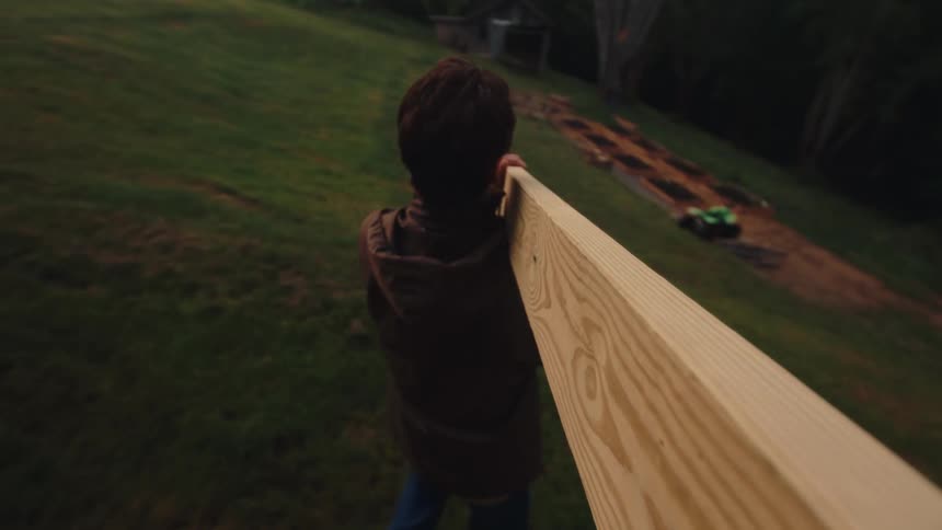 A boy helps with a construction project, carrying a heavy wooden plank across the yard in slow motion