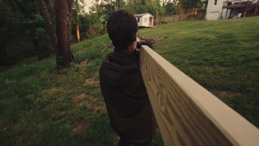 A young man emerges from a dark shed carrying a heavy wooden plank on his shoulder across a green yard during golden hour