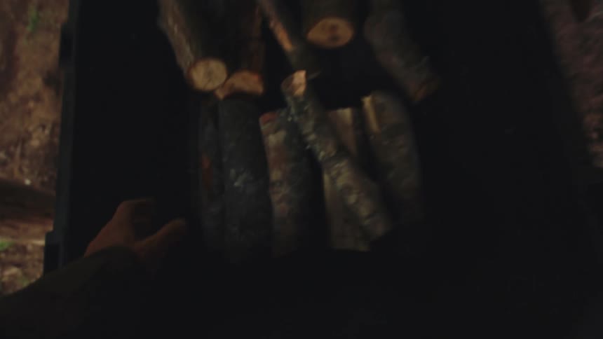 A first-person view of a man gathering firewood from a bin and stacking it neatly inside a rustic log cabin