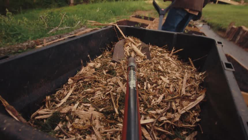 First-person perspective from a shovel in a wheelbarrow full of wood chips being moved through a garden with raised beds