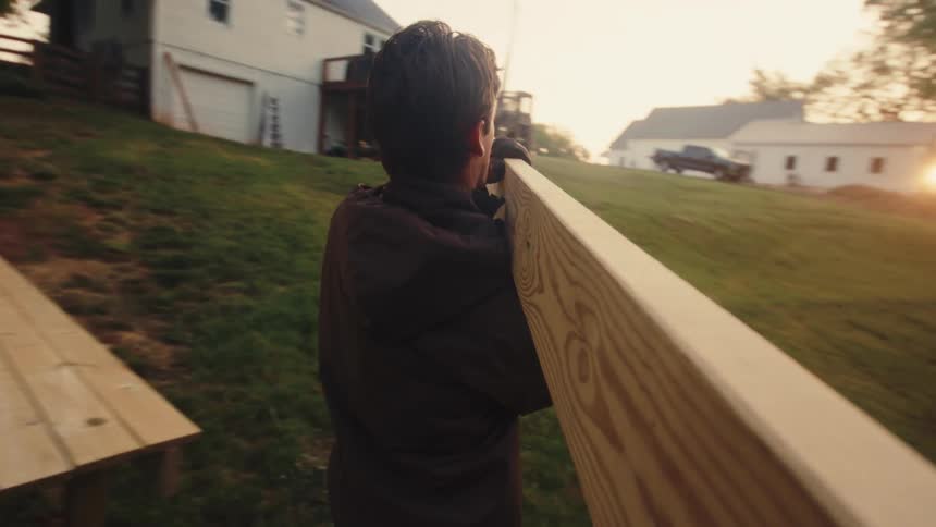 First-person view of a man carrying a long wooden plank across a grassy yard at golden hour in slow motion