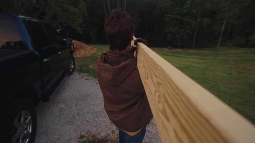 Point-of-view shot of a young man carrying a heavy wooden beam on his shoulder after unloading it from a pickup truck in a rural setting