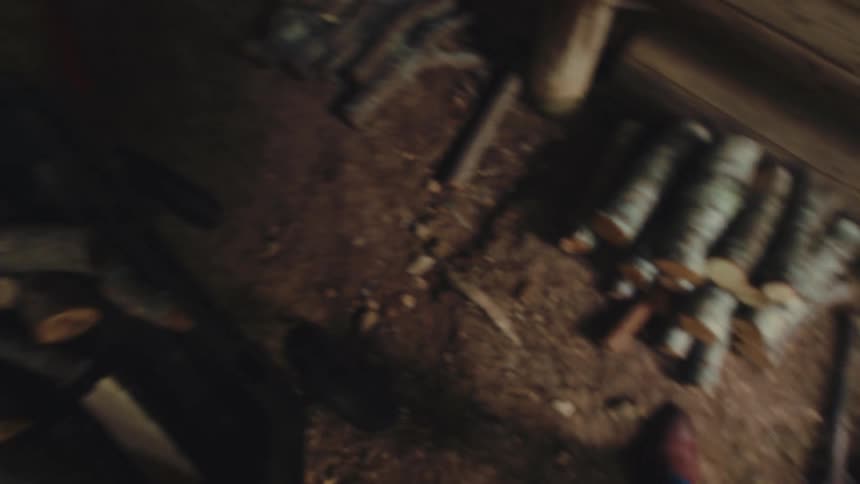 A first-person perspective of a man gathering chopped logs and placing them into a wheelbarrow inside a rustic shed