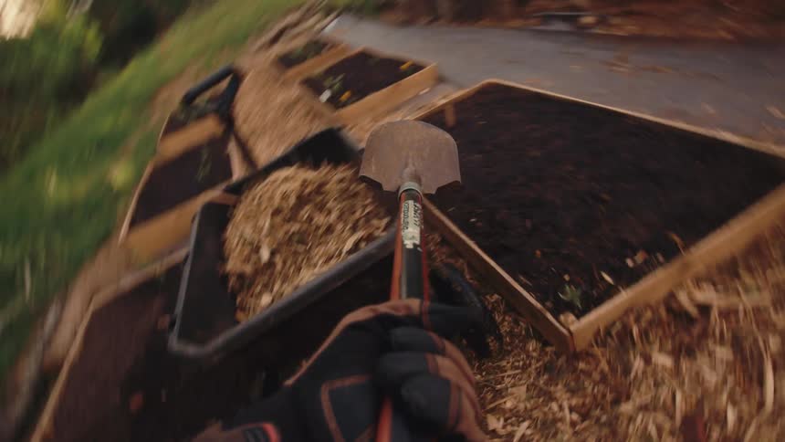 First-person perspective of a gardener shoveling and spreading wood chip mulch from a wheelbarrow onto the pathways of a backyard raised-bed garden