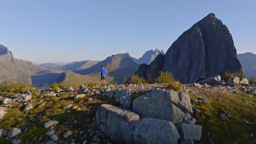 An aerial view reveals a man standing on a rocky mountain ridge overlooking a majestic fjord and peaks in the distance
