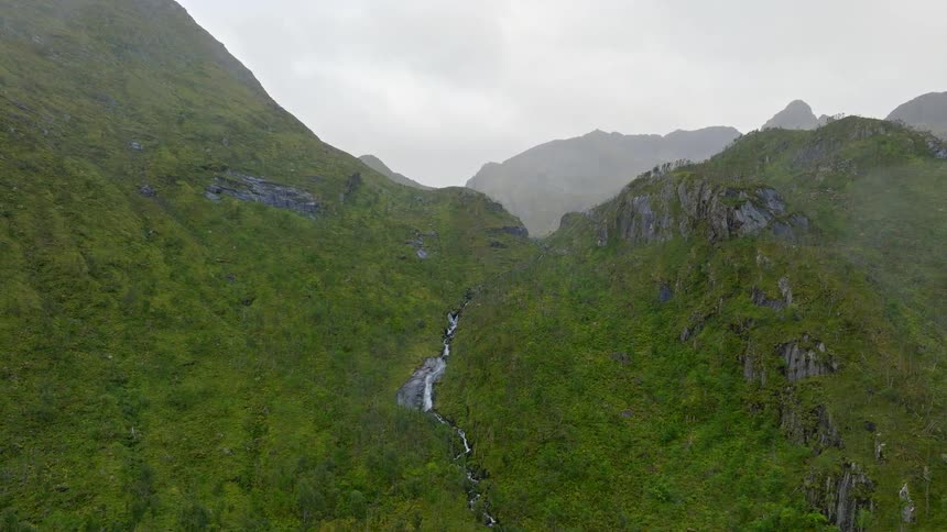 An aerial view shows a stunning waterfall cascading down a lush green mountain on a foggy day