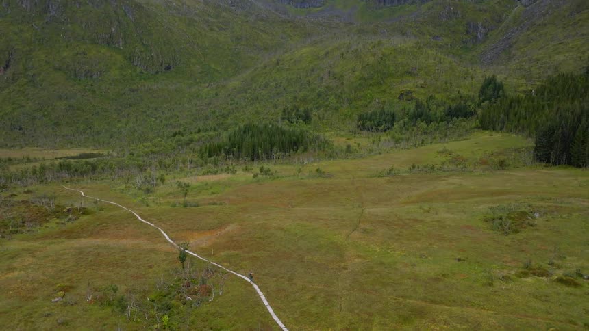 Aerial view of a lone hiker walking along a wooden boardwalk through a vast green moorland towards a majestic mountain range