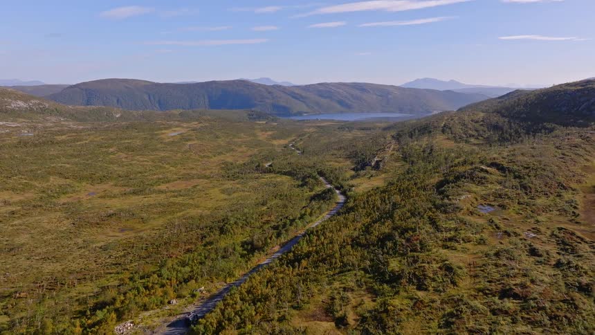 Aerial footage follows a car driving on a winding road through a vast, green mountain valley towards a distant lake