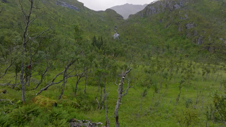 Aerial shot pushes past a solo hiker relaxing at a remote campsite, revealing a magnificent view of a waterfall cascading down a verdant mountain slope