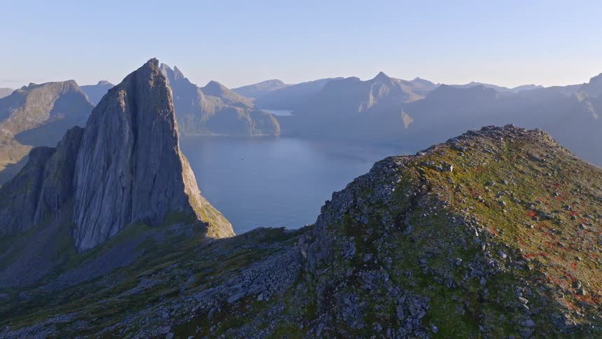 A breathtaking aerial shot pulls back along a rocky ridge to reveal the iconic, jagged mountains and calm fjords of Senja, Norway
