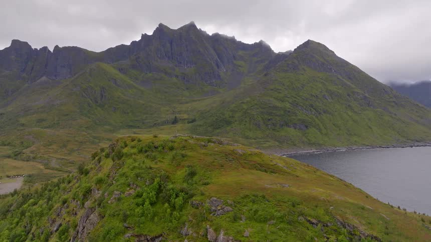 An epic aerial orbit of a lush green bluff reveals a rugged coastline and jagged mountains in Norway