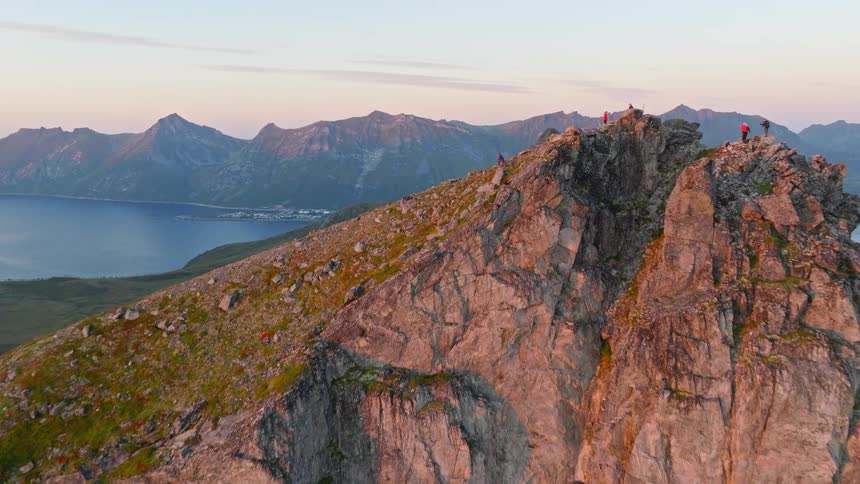 Aerial drone shot flying over a dramatic, rocky mountain peak where hikers enjoy the beautiful sunset view
