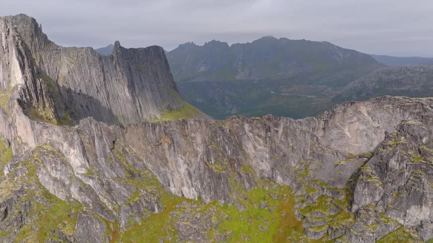 An epic aerial reveal of a vast, rocky mountain range with towering peaks overlooking a deep valley and the distant sea