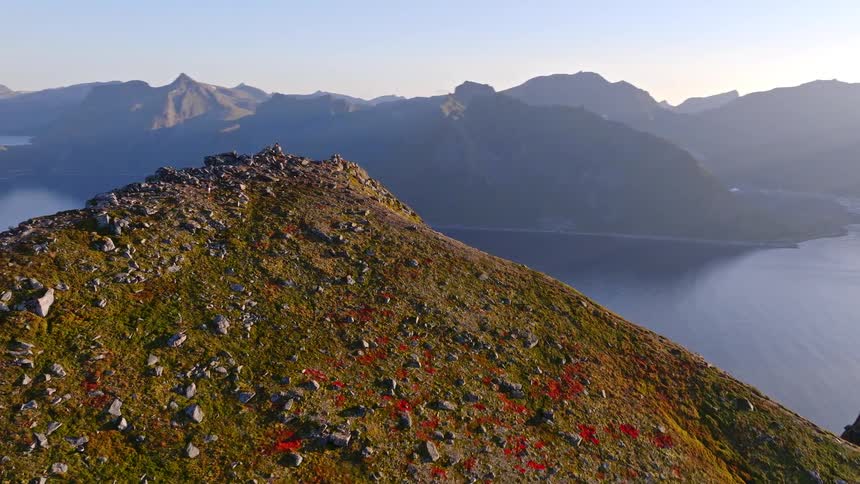An epic aerial shot ascends over a rugged mountain ridge, revealing a breathtaking fjord and majestic peaks under the warm morning sun