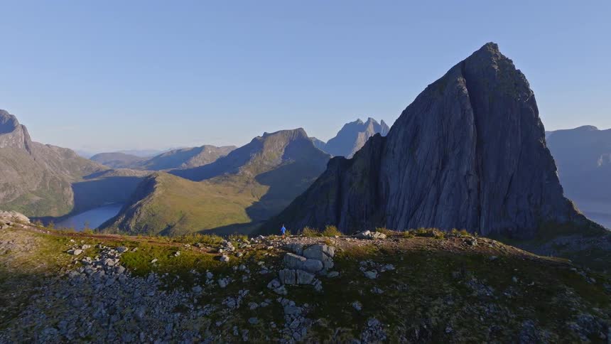 Aerial drone shot pulls back to reveal a lone hiker on a majestic mountain ridge overlooking a stunning fjord valley during golden hour