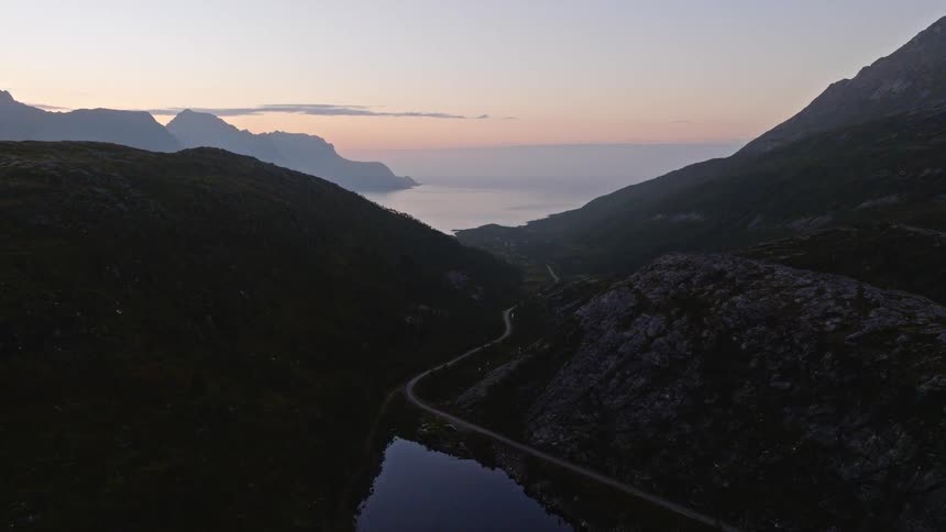 Static aerial view of a tranquil lake and a winding road through a mountain valley leading to the sea at dusk