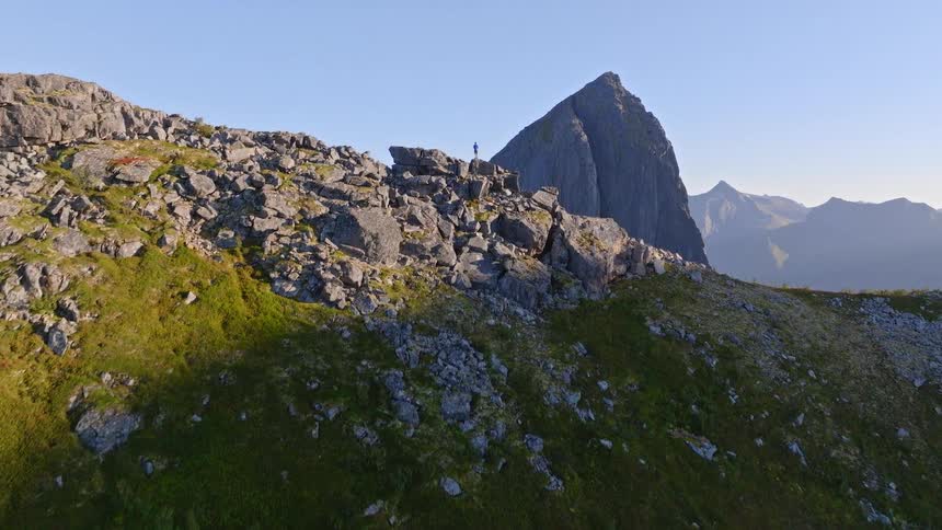 A stunning aerial shot reveals a lone hiker standing on a rocky mountain ridge, showcasing the breathtaking scale of the majestic peaks in the background