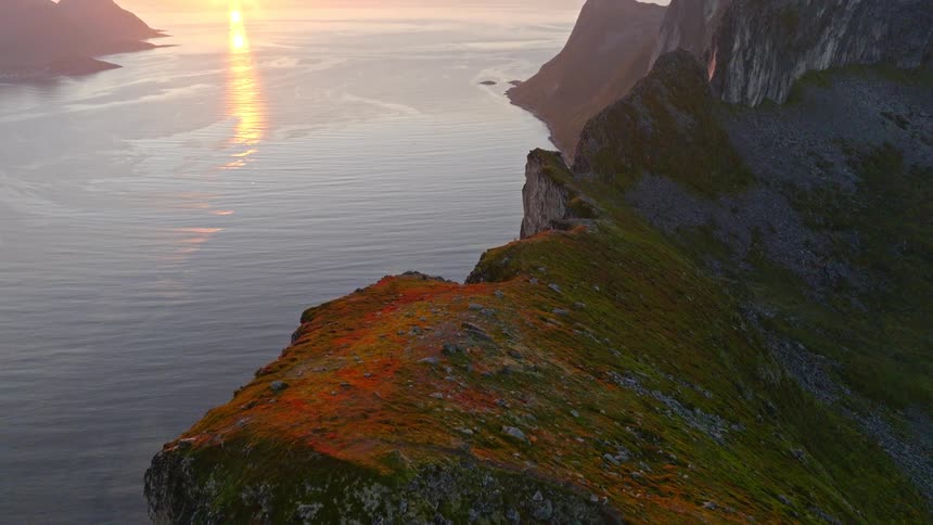 An aerial shot flies along a vibrant, colorful mountain cliffside, revealing a spectacular sunset over a majestic ocean fjord