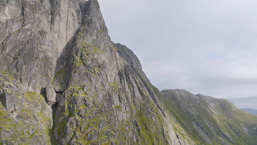 A majestic aerial view slowly ascends the steep, rugged rock face of a massive mountain covered in patches of green moss