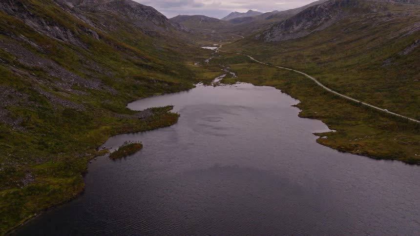 A dramatic aerial view pans up over a dark lake to reveal a vast, remote mountain valley under an overcast sky