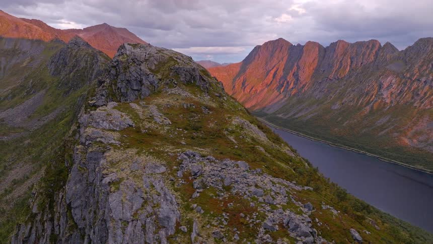 A stunning aerial view pulls back from a rocky mountain ridge to reveal a majestic valley with a serene lake, as distant peaks glow in the golden light of sunset