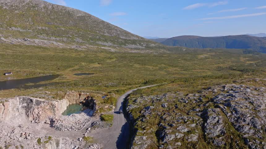 An aerial drone follows a car driving on a remote road through a vast, rocky mountain landscape past an old quarry
