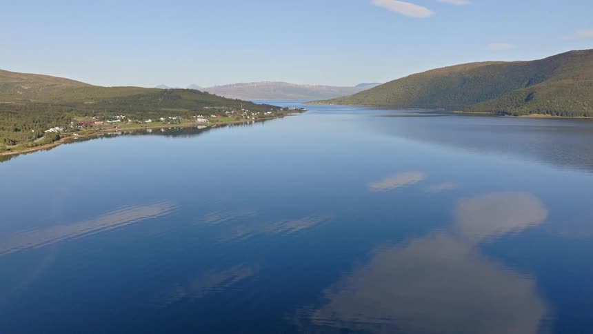 An expansive aerial shot slowly retreats over a pristine, calm blue lake in a mountainous landscape, showing a small coastal village and perfect sky reflections