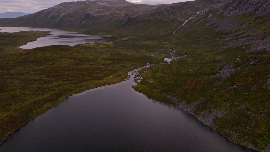 A moody aerial shot tilts up over a dark, still lake to reveal a vast, remote mountain valley under a cloudy sky
