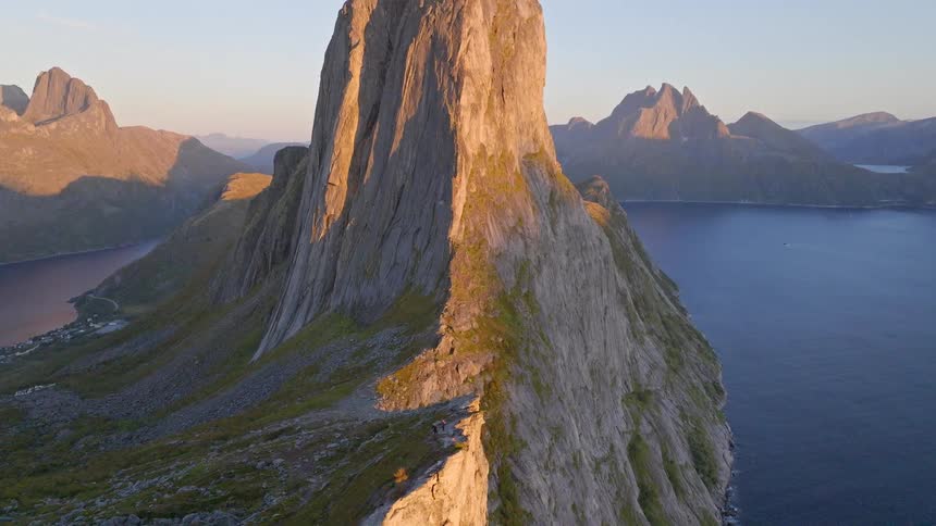 A stunning aerial view pulls back to reveal campers on a sheer cliff edge of a massive mountain surrounded by fjords at sunset in Norway