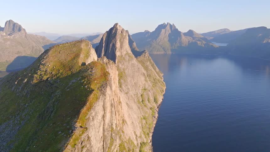 A majestic aerial view slowly reveals the dramatic, sunlit ridges of the Segla mountain in Senja, Norway, towering over the calm blue fjord