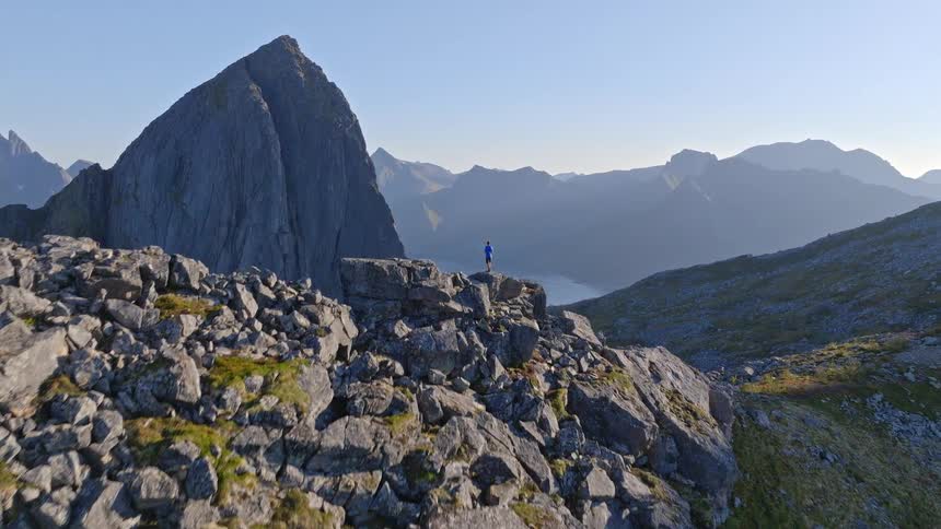 An aerial view slowly reveals an epic mountain landscape as it pushes in on a lone hiker standing on a rocky precipice at sunrise