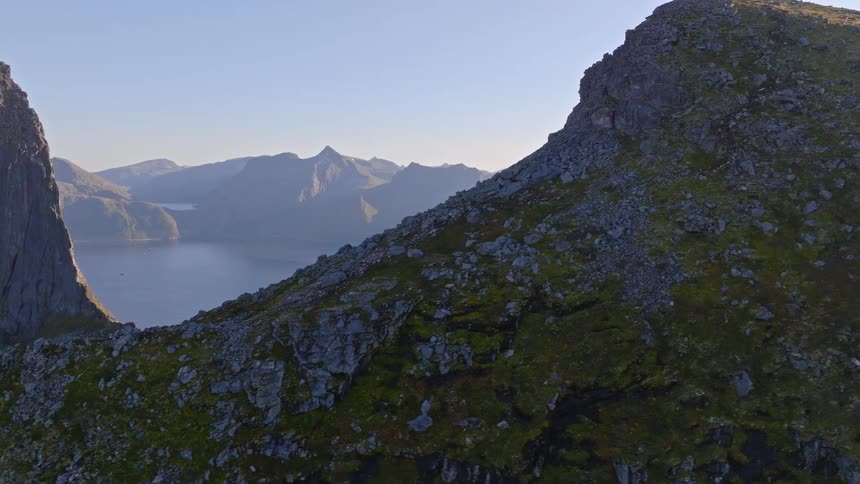 An awe-inspiring aerial view reveals a vast, rugged mountain landscape and a fjord as the camera pulls back along a rocky ridge towards the summit