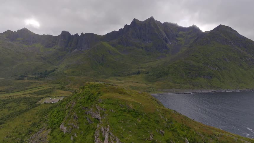 A slow aerial pullback reveals a vast and rugged green mountain range meeting the ocean on a cloudy day in Norway