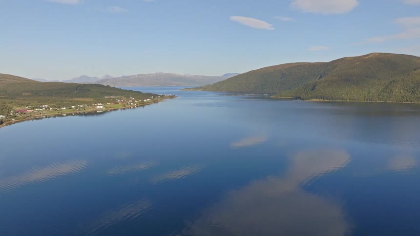 A slow aerial tilt-up reveals a serene Scandinavian fjord with glassy water reflecting a clear blue sky