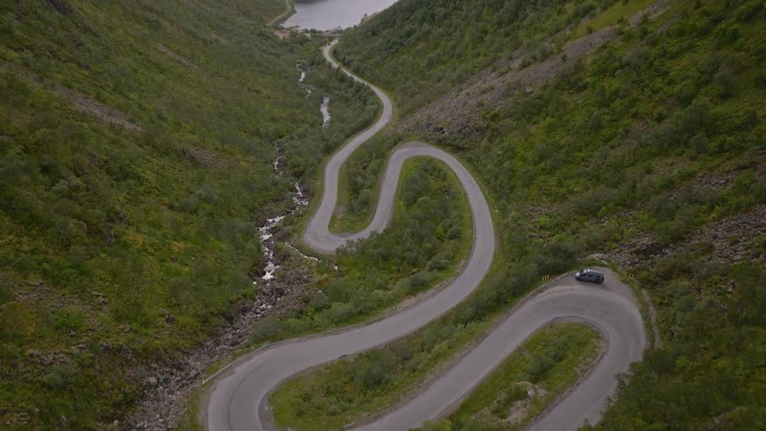 An aerial shot pulls back to reveal a long, winding road snaking through a lush green mountain valley towards a distant fjord