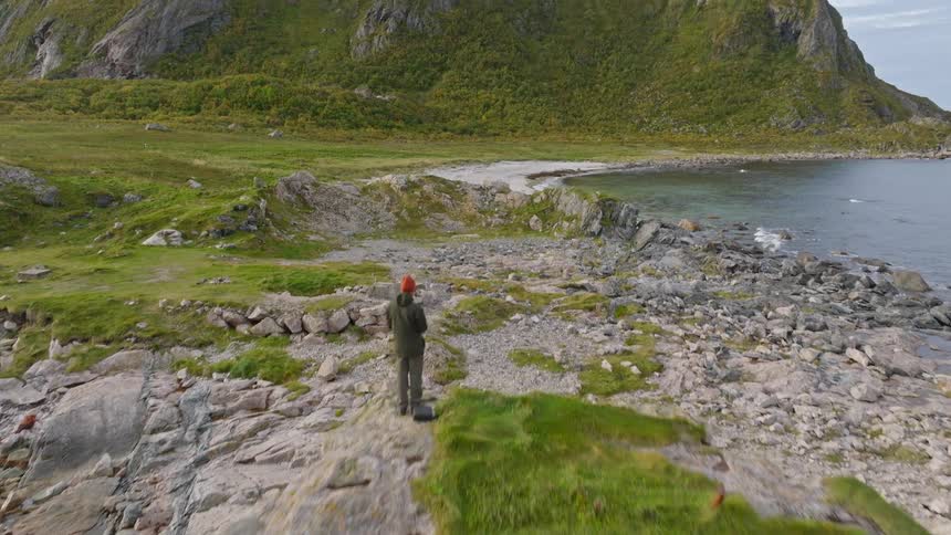 Aerial footage captures a hiker on a rocky outcrop overlooking a stunning Norwegian coastline with a mountain and beach
