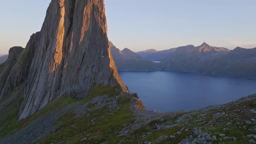 A majestic, sun-kissed mountain peak towers over a deep blue fjord during a serene golden hour