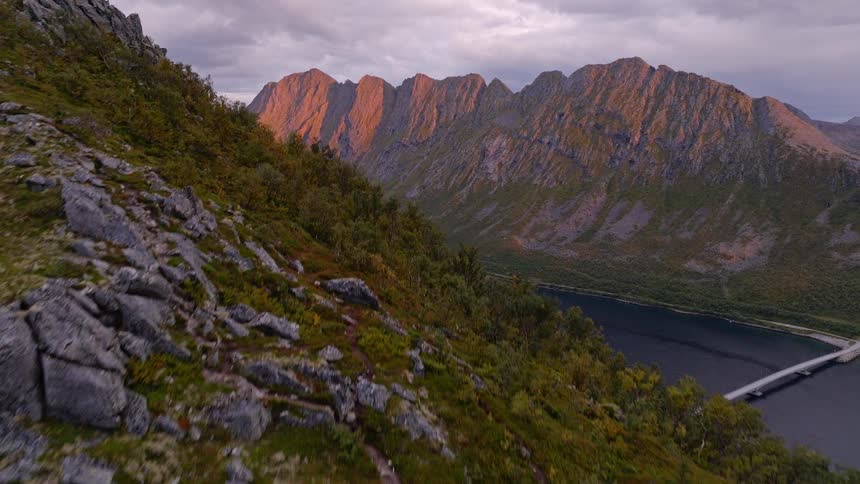Aerial shot rises over a rugged, rocky cliff to reveal a breathtaking view of sun-kissed mountains and a tranquil fjord at sunset