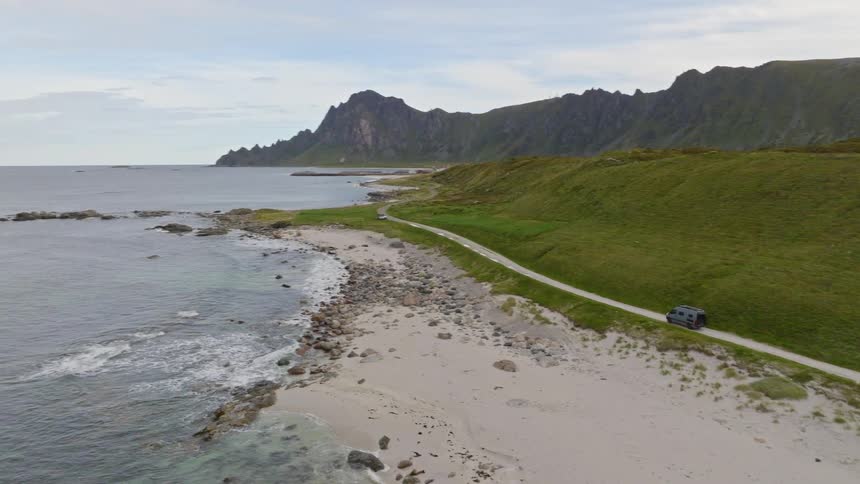 A stunning aerial shot follows a camper van driving along a remote coastal road between a white sand beach and verdant hills